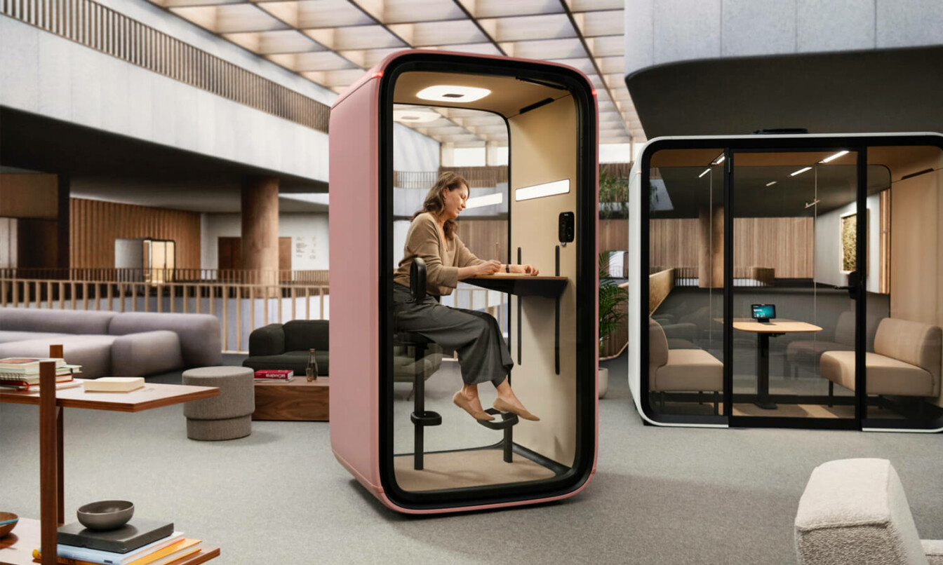 A woman sitting inside a blush color Framery phone booth in a modern Scandinavian office setting.