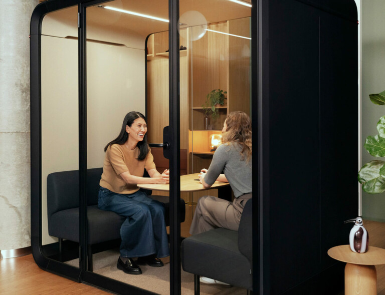 Two women sitting inside a black framed Framery meeting pod, the other woman facing the camera.