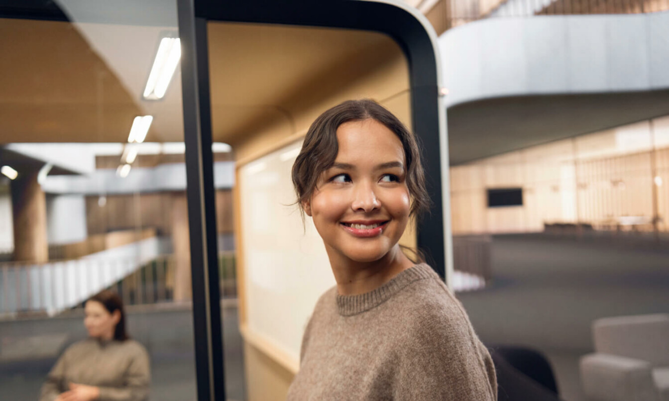 A dark haired woman with a camel colored shirt smiling towards and entering a Framery meeting pod.