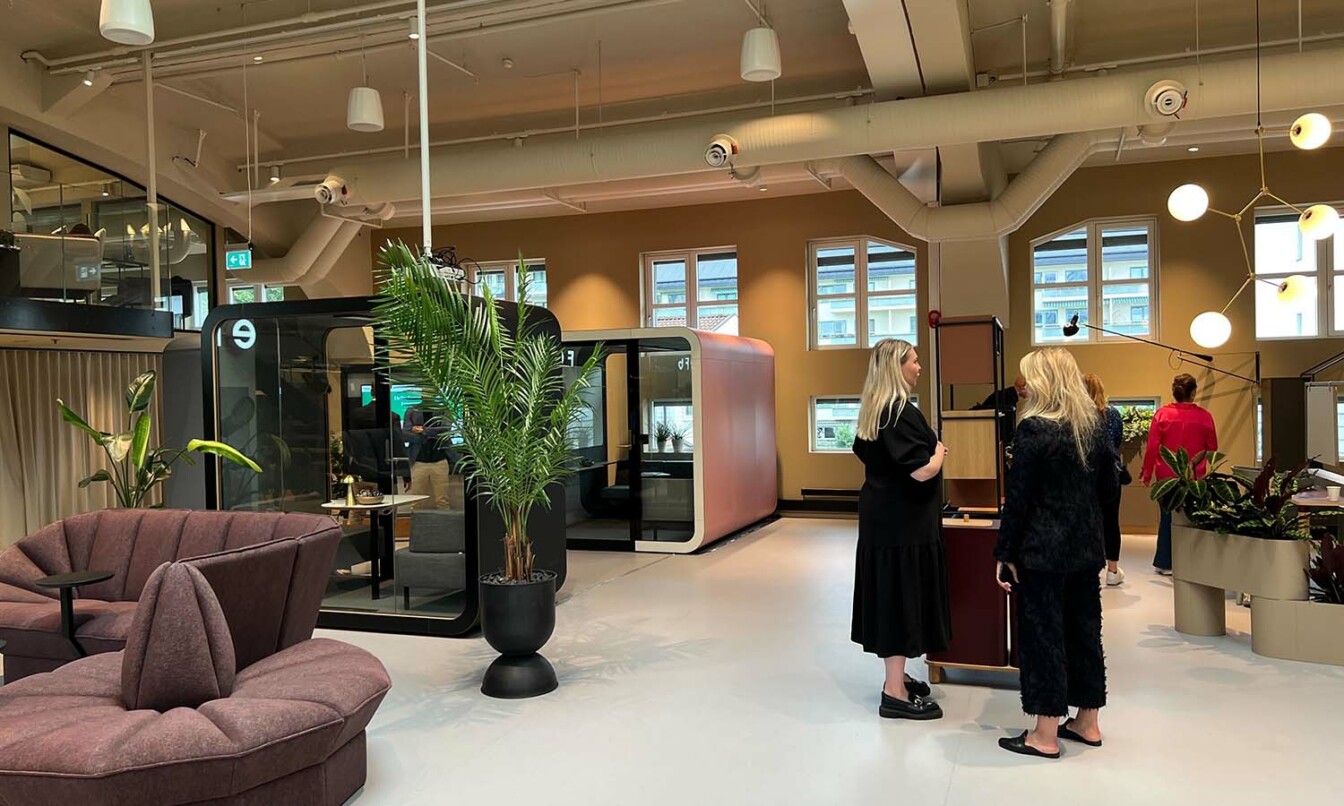 People having a conversation in an open space containing Framery 2Q meeting room and a Framery Q meeting pod.