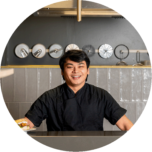 A smiling person in chef clothes standing in front of a kitchen.