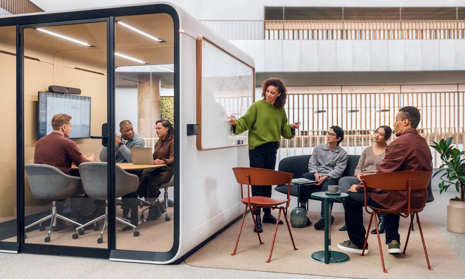 A group of people having a meeting next to a soundproof meeting space, with people inside the meeting space as well.