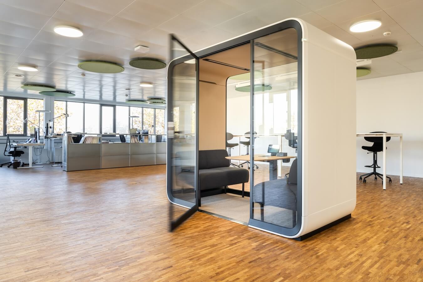 An office setup with a beige meeting pod at the front with its door open.