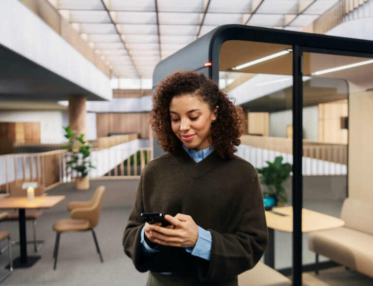 Woman looking at her phone and standing in front of a meeting pod in a scandinavia style office.