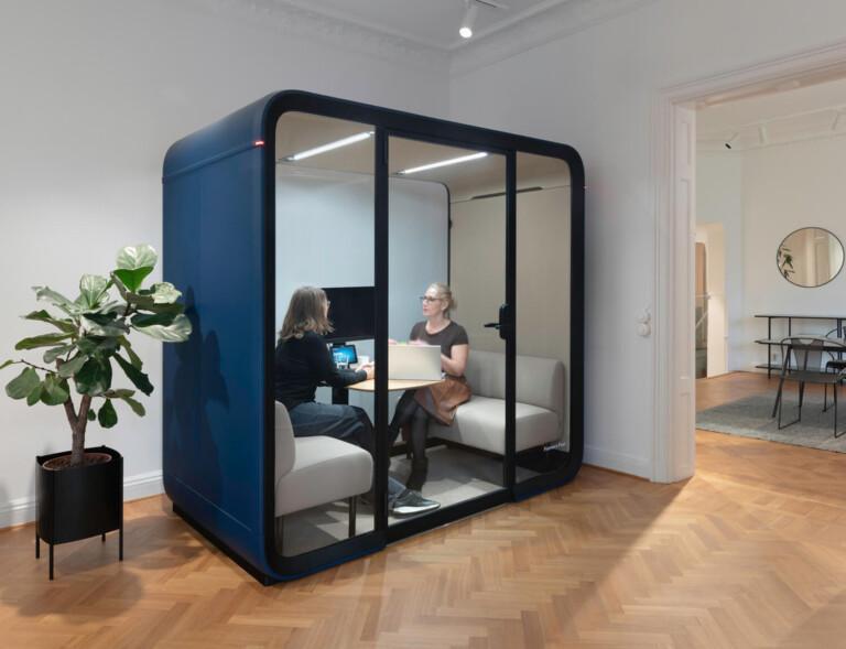 Two women sitting inside a navy blue Framery Four office pod.