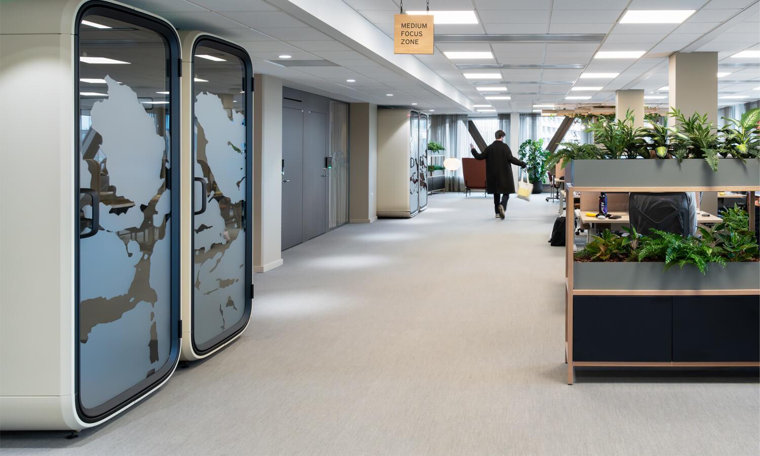 A wide corridor in an open office, with acoustic pods on the other side and desks on the other.