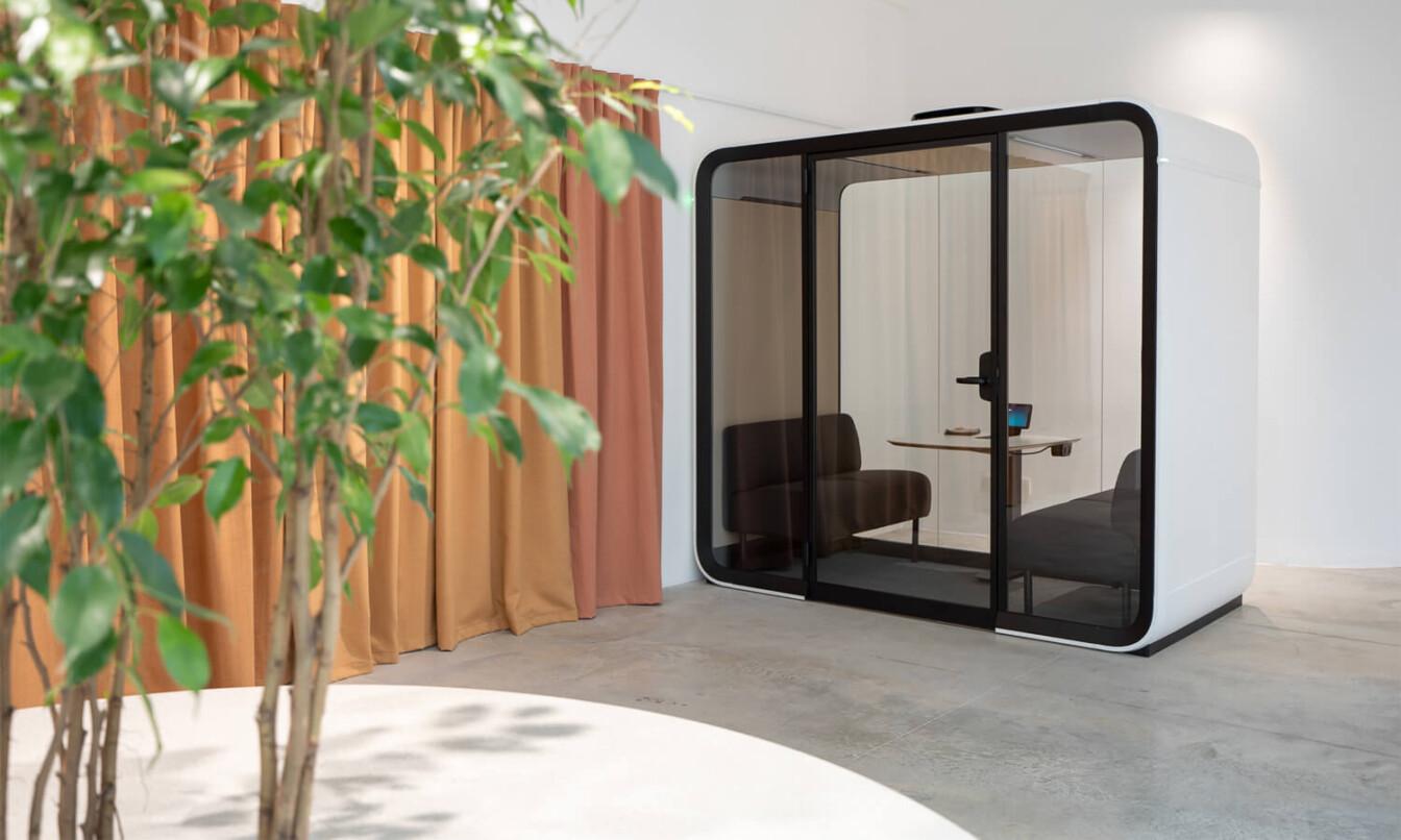 A white Framery Four meeting pod next to an cinnamon colored curtain and a green tree.