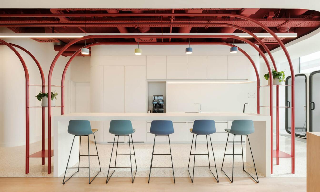 A light office kitchen area with a red metal structure surrounding table with colorful chairs.