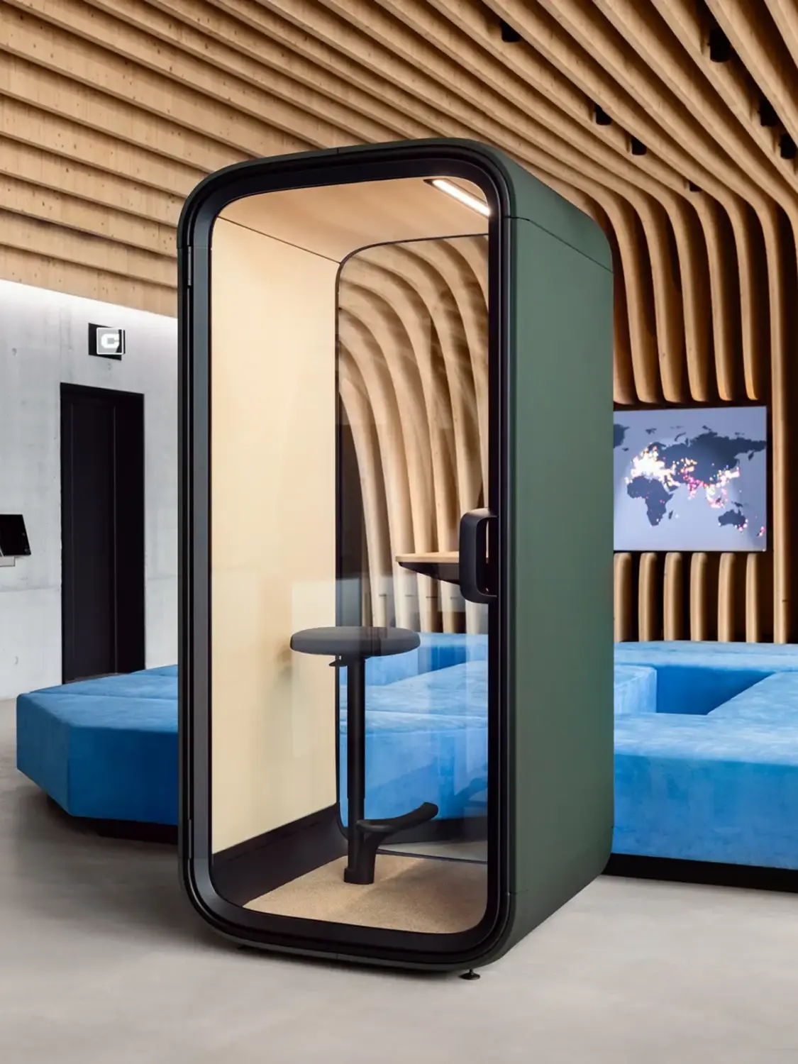 Modern office space with a sleek Framery One Compact phone booth, curved wooden ceiling, blue seating, and a world map screen on the wall.