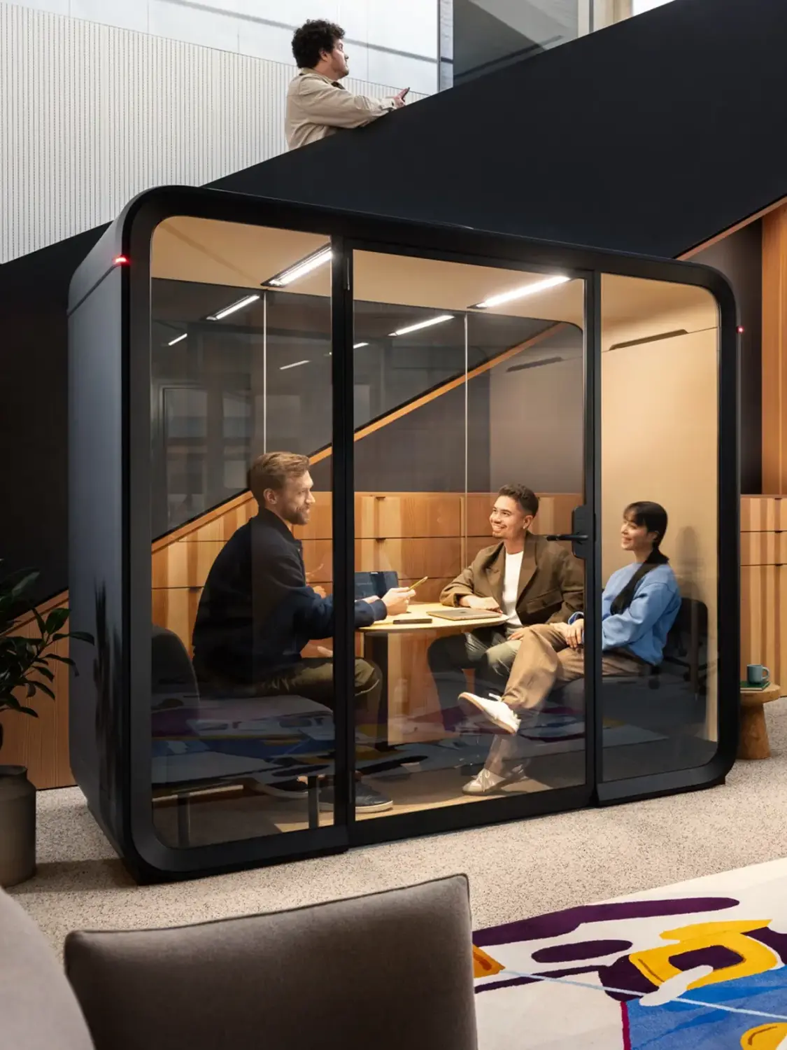 Three people converse inside a Framery Four meeting pod, while another person stands outside. The setting appears modern and contemporary.