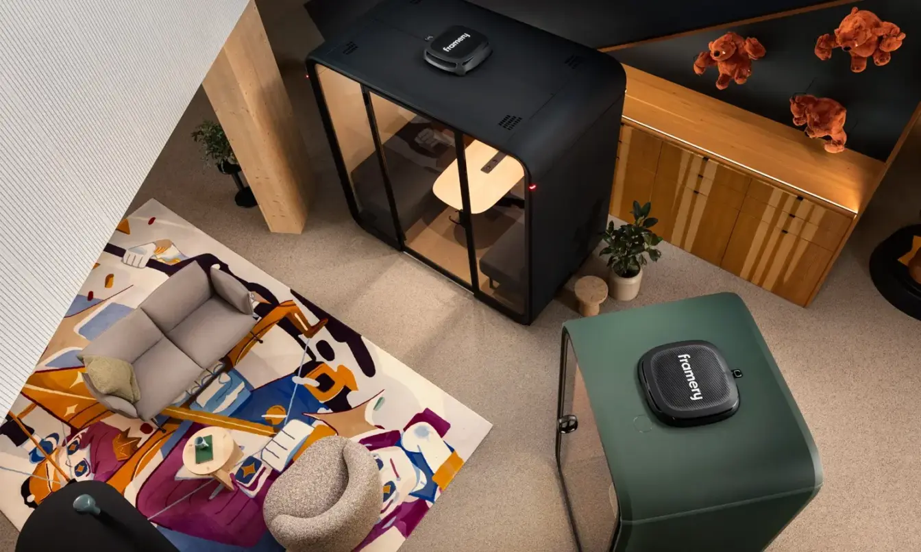 Modern office space with soundproof Framery pods, colorful abstract rug, and couches. Features contemporary furniture and decor with potted plants and playful stuffed animals.