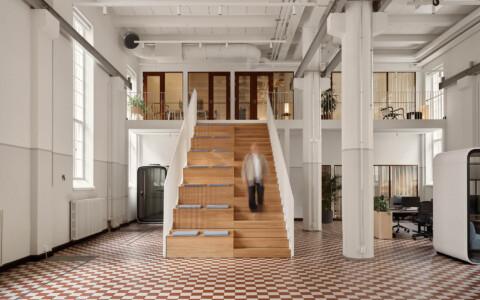Spacious, modern office interior with a person descending wooden stairs. Features open workspaces, Framery smart pods, checkered floor, and white walls with large windows.