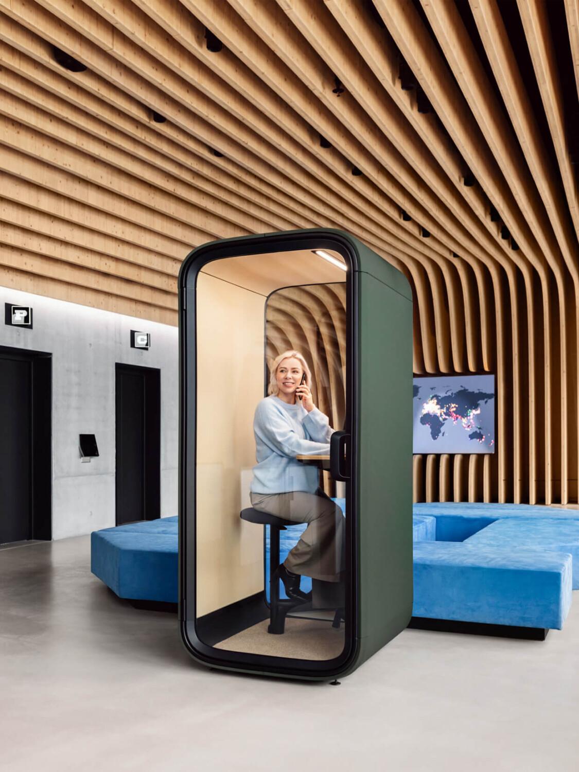 A person sits in a modern Framery phone booth within a stylish office space, featuring wooden ceiling panels and a blue couch.