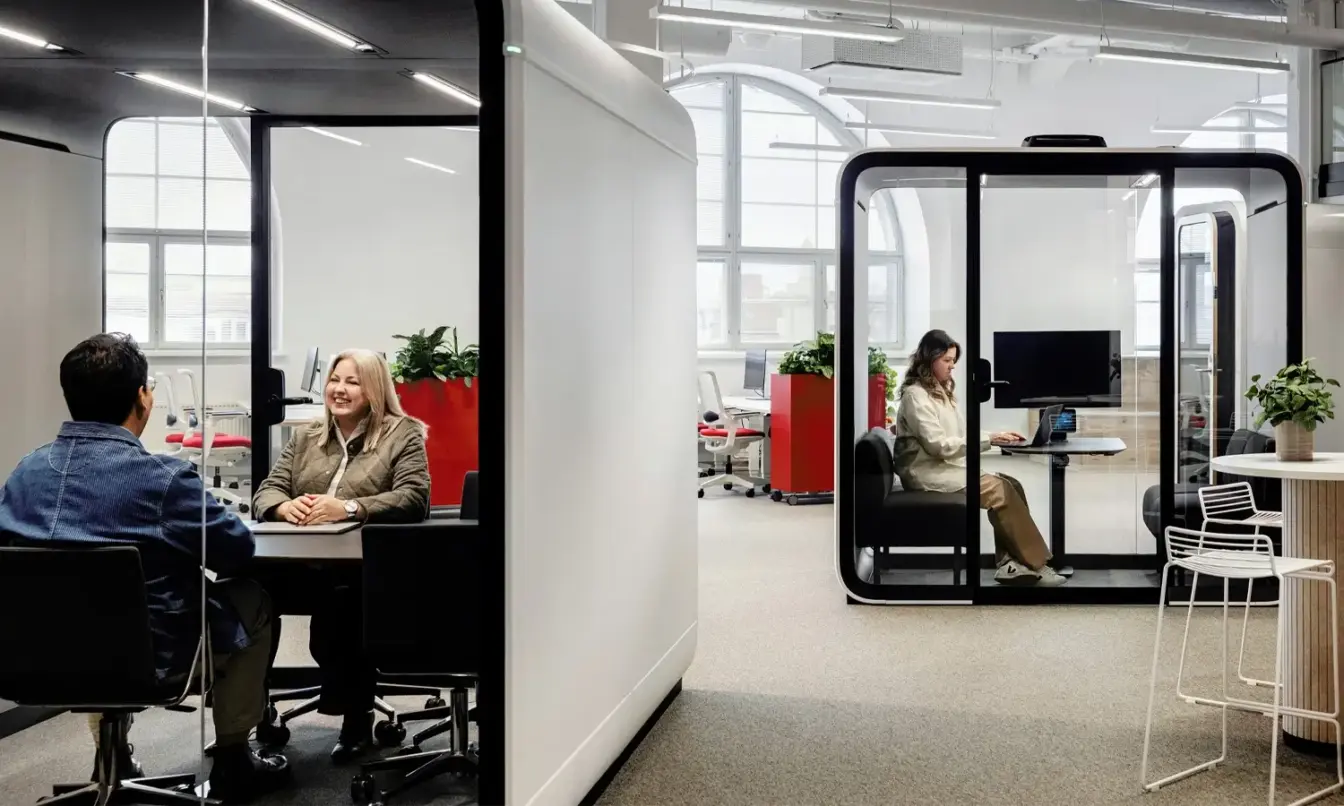 Office space with people in Framery Six and Four meeting pods, working on computers. Red accents and potted plants enhance the minimalist, collaborative environment. Bright, spacious design.