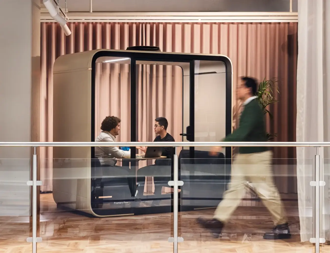 Two people sit across from each other inside a glass-walled Framery Four meeting pod as a blurred passerby walks by in the foreground.
