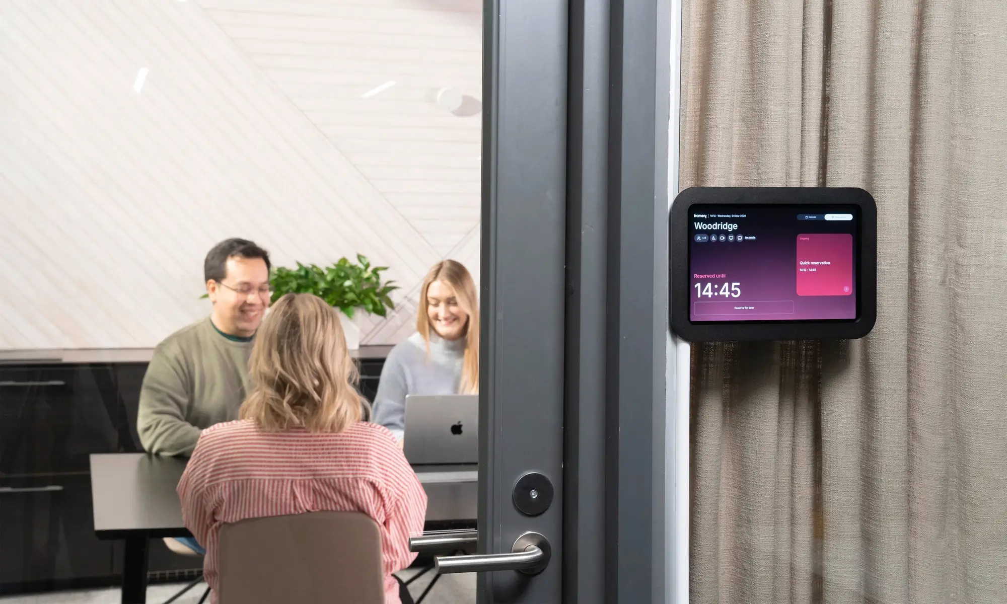 A small group of people sits around a table in a bright meeting room, with one facing away from the camera and a wall-mounted smart display nearby.