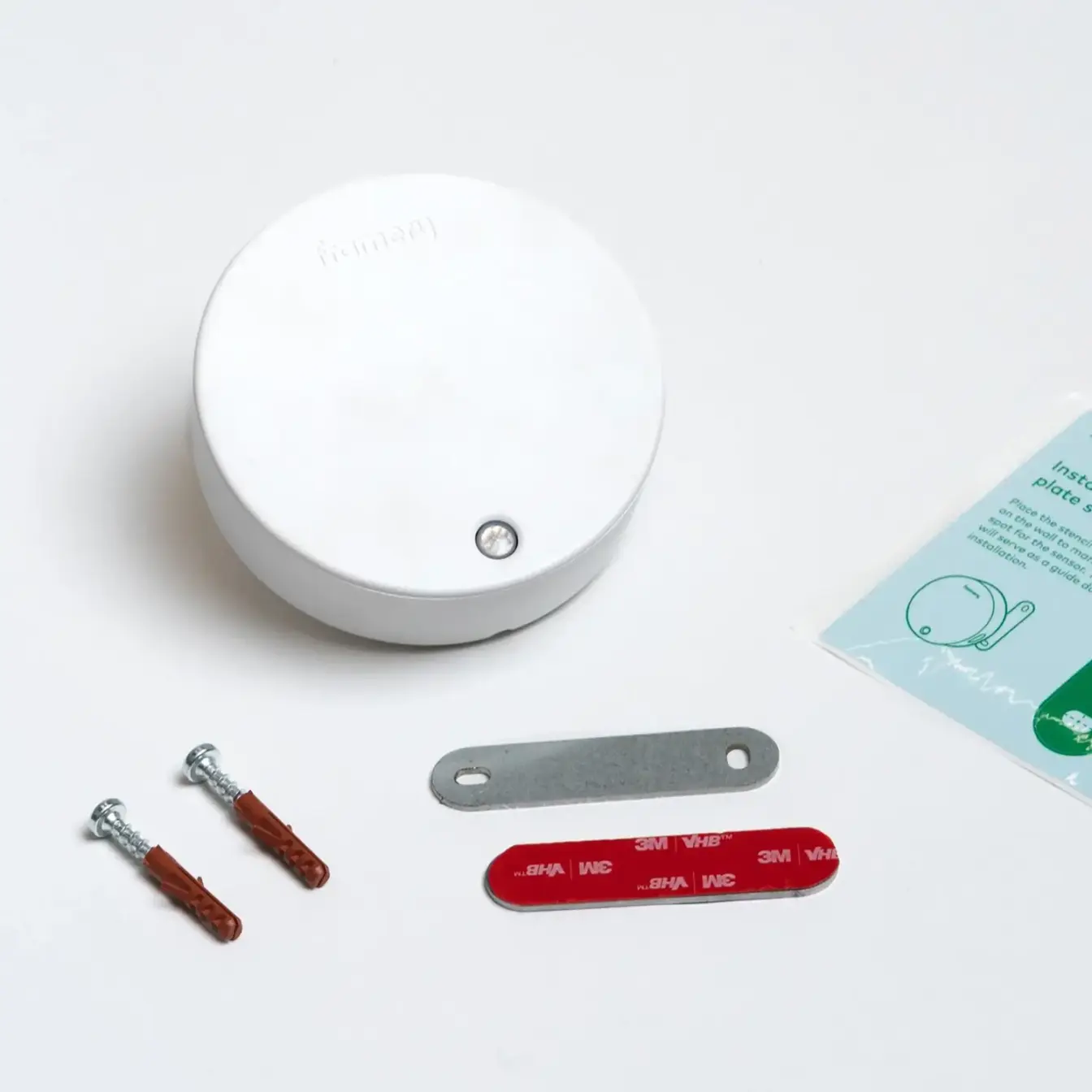 Un dispositivo Framery Room Sensor circular de color blanco con pequeñas piezas de ferretería -dos tornillos, un soporte metálico, una tira roja- y una tarjeta de instrucciones sobre una superficie blanca.
