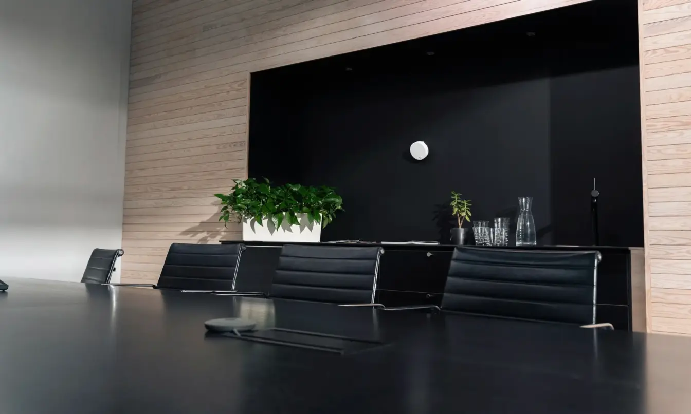 Modern conference room with a long dark table, black leather chairs, a potted plant, and glassware on a counter against a wood-paneled wall.