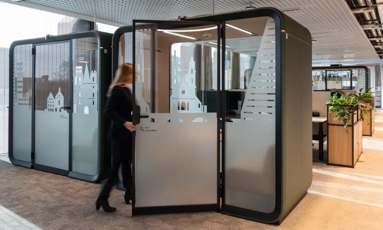 A person walks into a glass-walled meeting pod in a modern office interior.