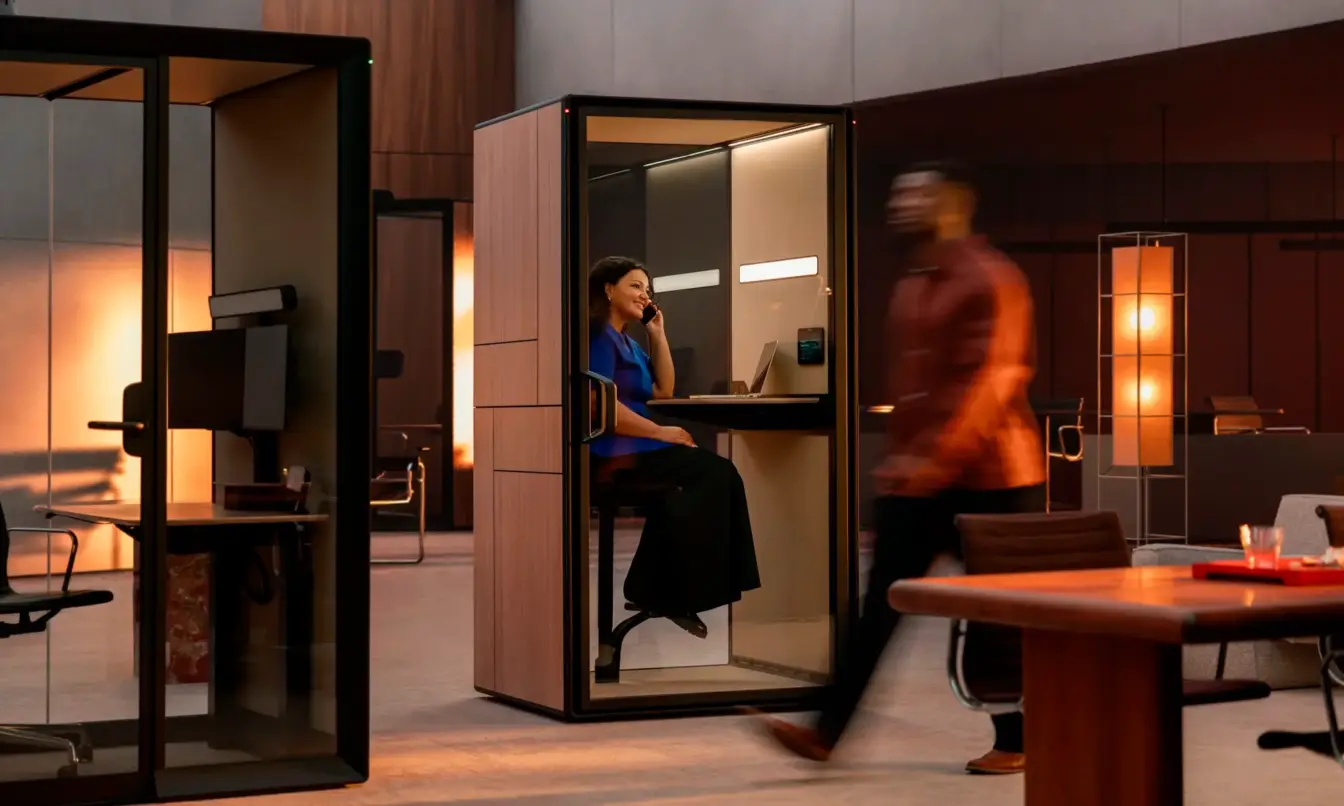 Modern office space with glass-walled Framery Gradus work pods; a person sits at a desk inside one booth while another person walks past in the foreground.