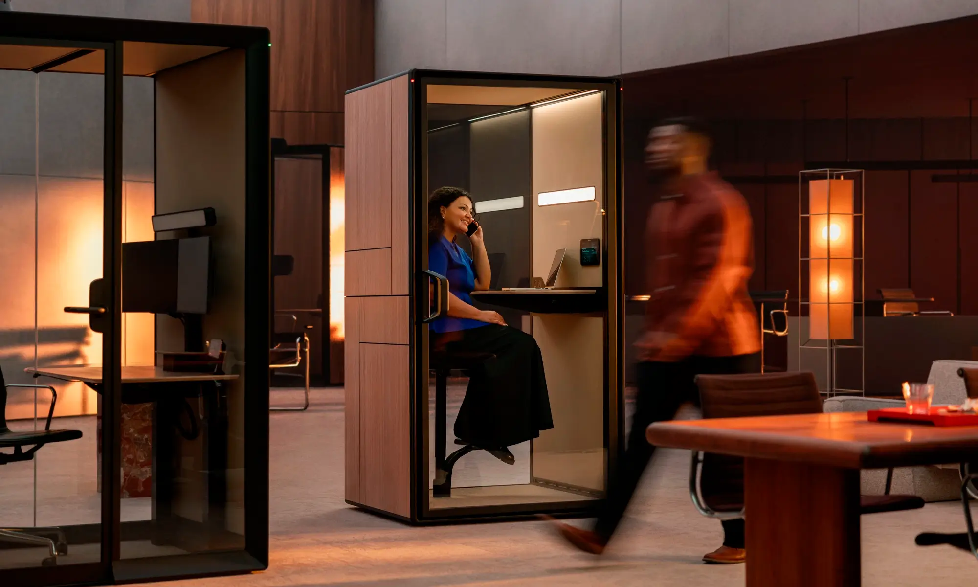 Modern office space with glass-walled Framery Gradus work pods; a person sits at a desk inside one booth while another person walks past in the foreground.