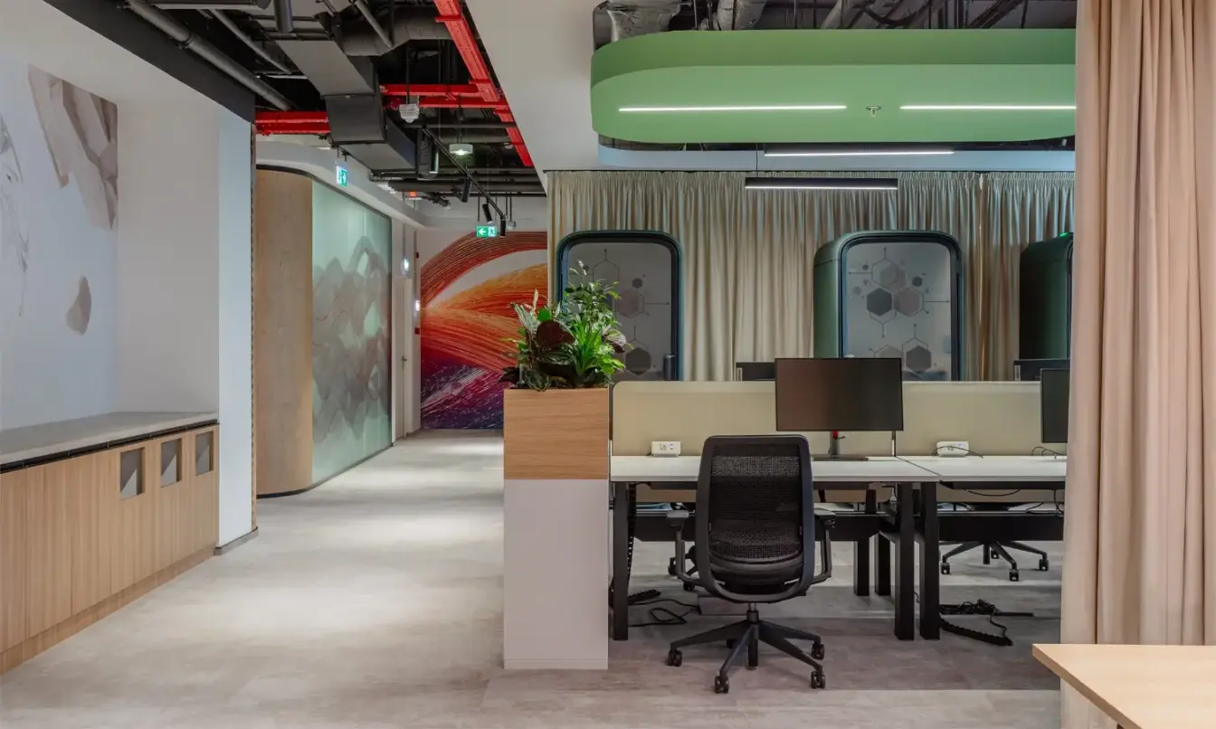 Open-plan office space with desks, chairs, Framery One Compact smart pods, and a potted plant on a divider.