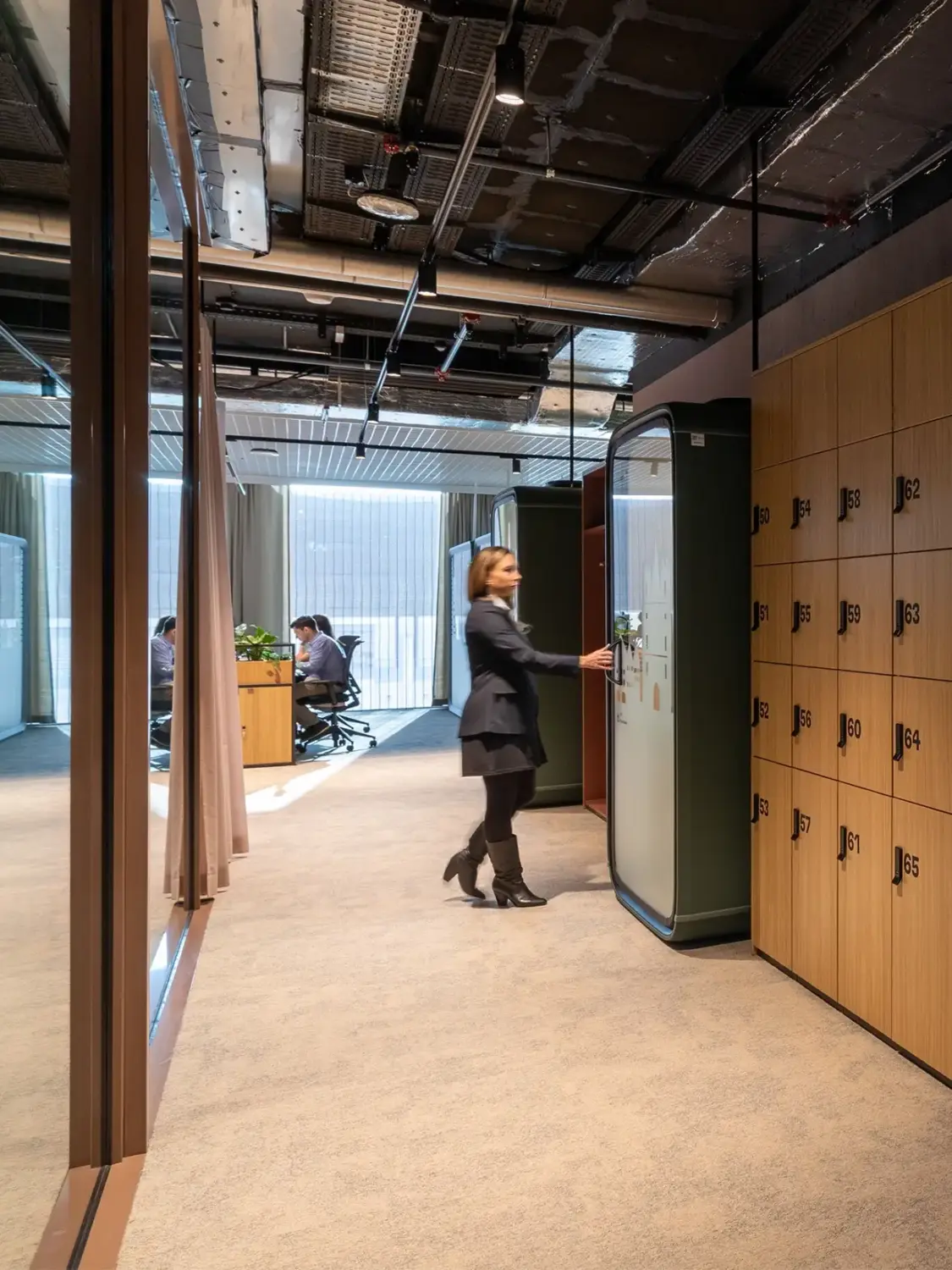 A businesswoman in a blazer opens the door of a Framery One smart pod in a modern office hallway, with wooden lockers along the right and coworkers at desks in the background.