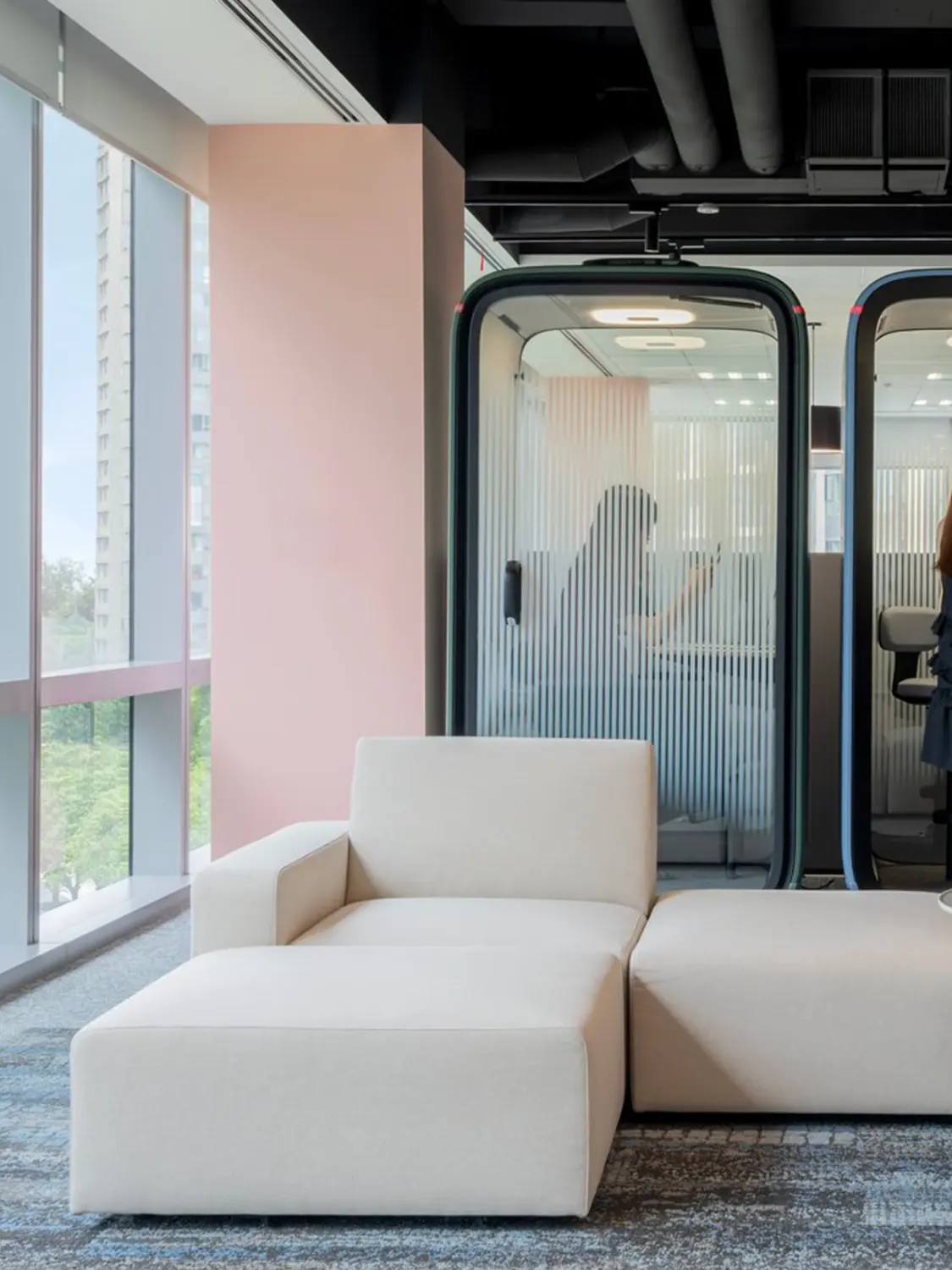 Modern office lounge featuring white modular seating, a pink column, and glass-walled meeting rooms.