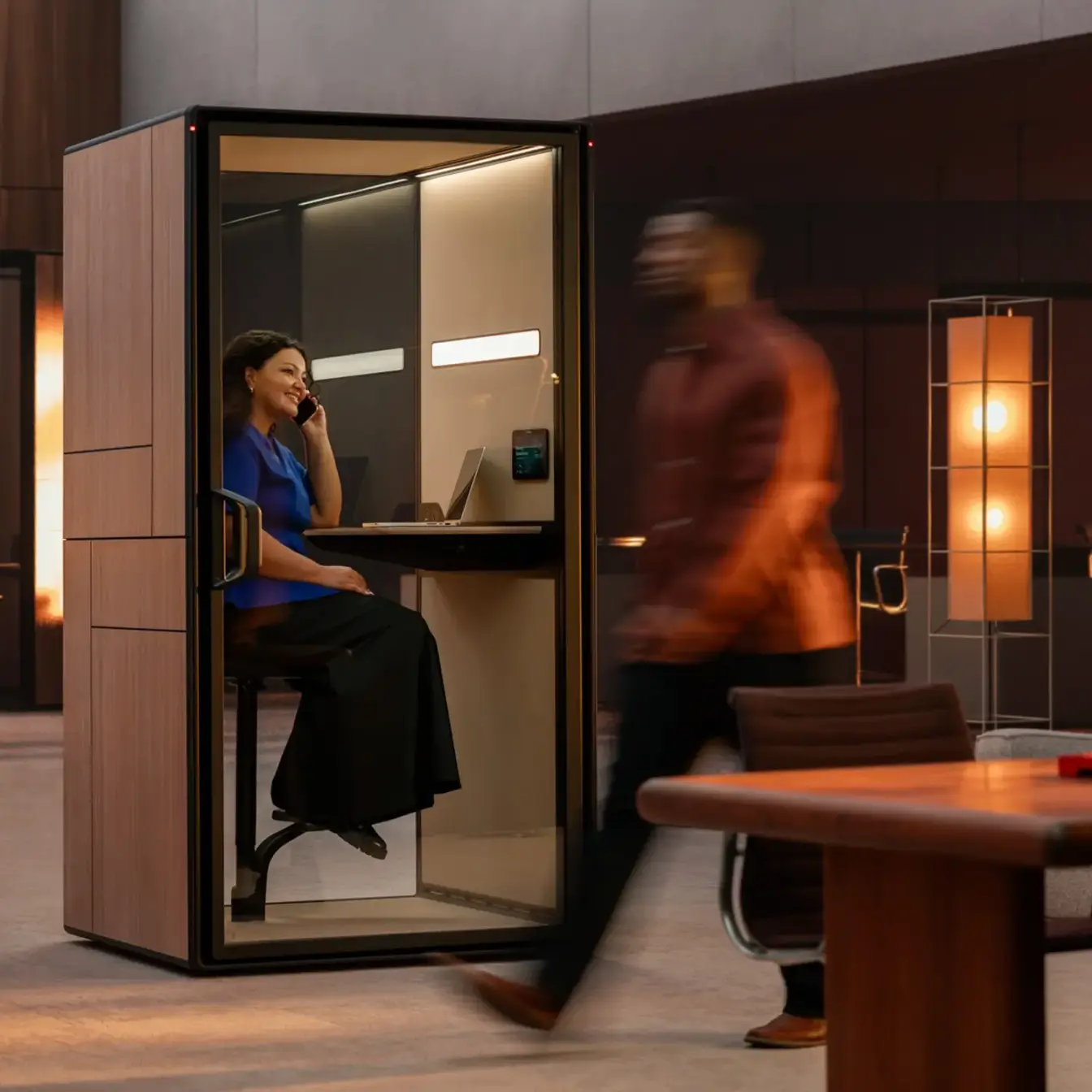 A woman sits inside a private Framery Gradus Focus booth talking on a phone, while a blurred man walks past in a modern lounge area.