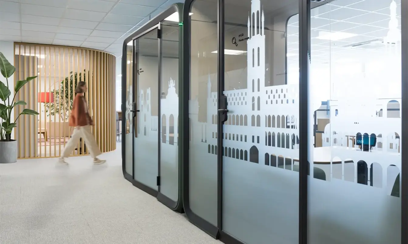 A modern office hallway with glass partition walls and a person walking in the background.