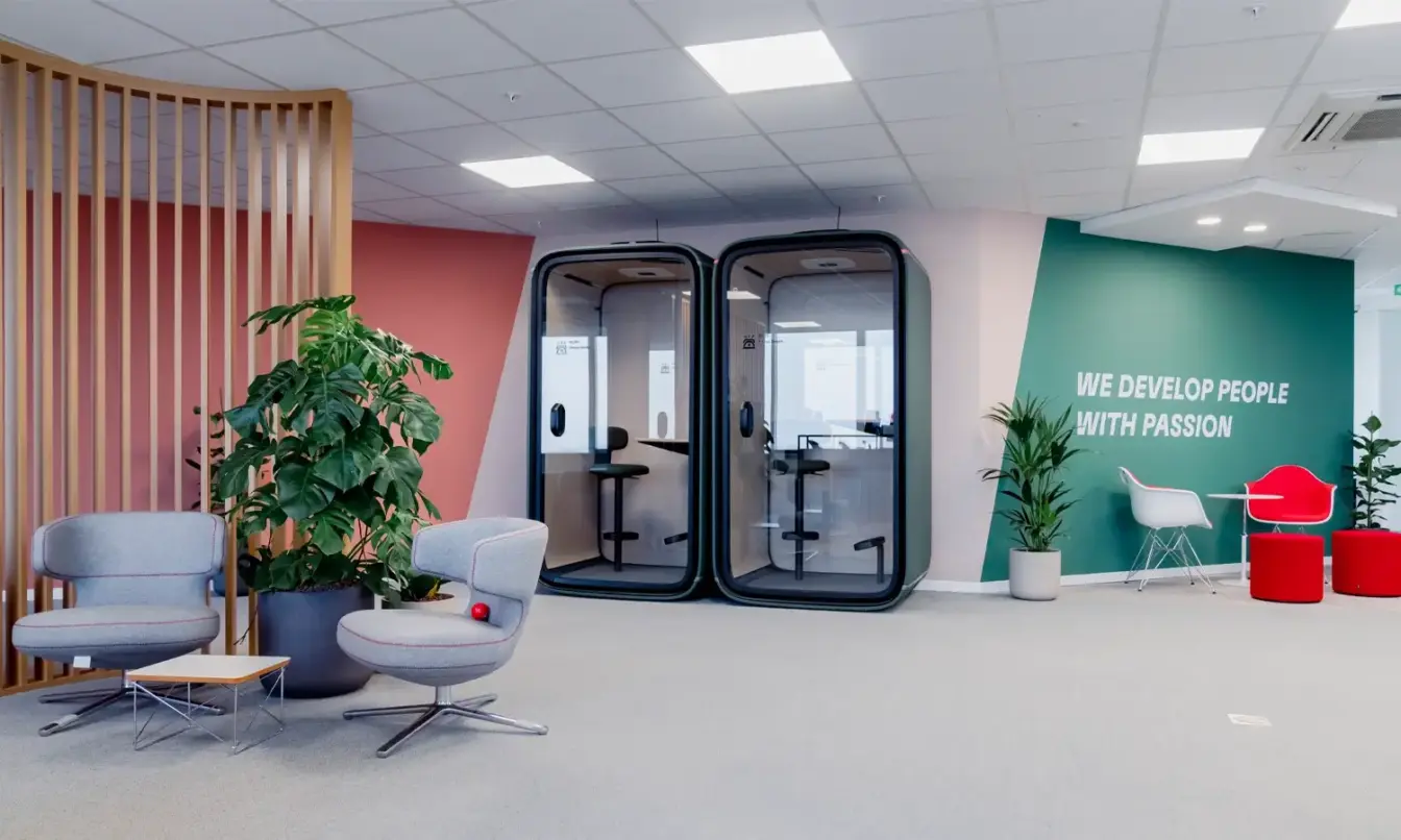Modern office lounge featuring glass double doors, a wooden slat divider with gray seating on the left, and a green accent wall with the phrase “WE DEVELOP PEOPLE WITH PURPOSE” and red stools on the right.