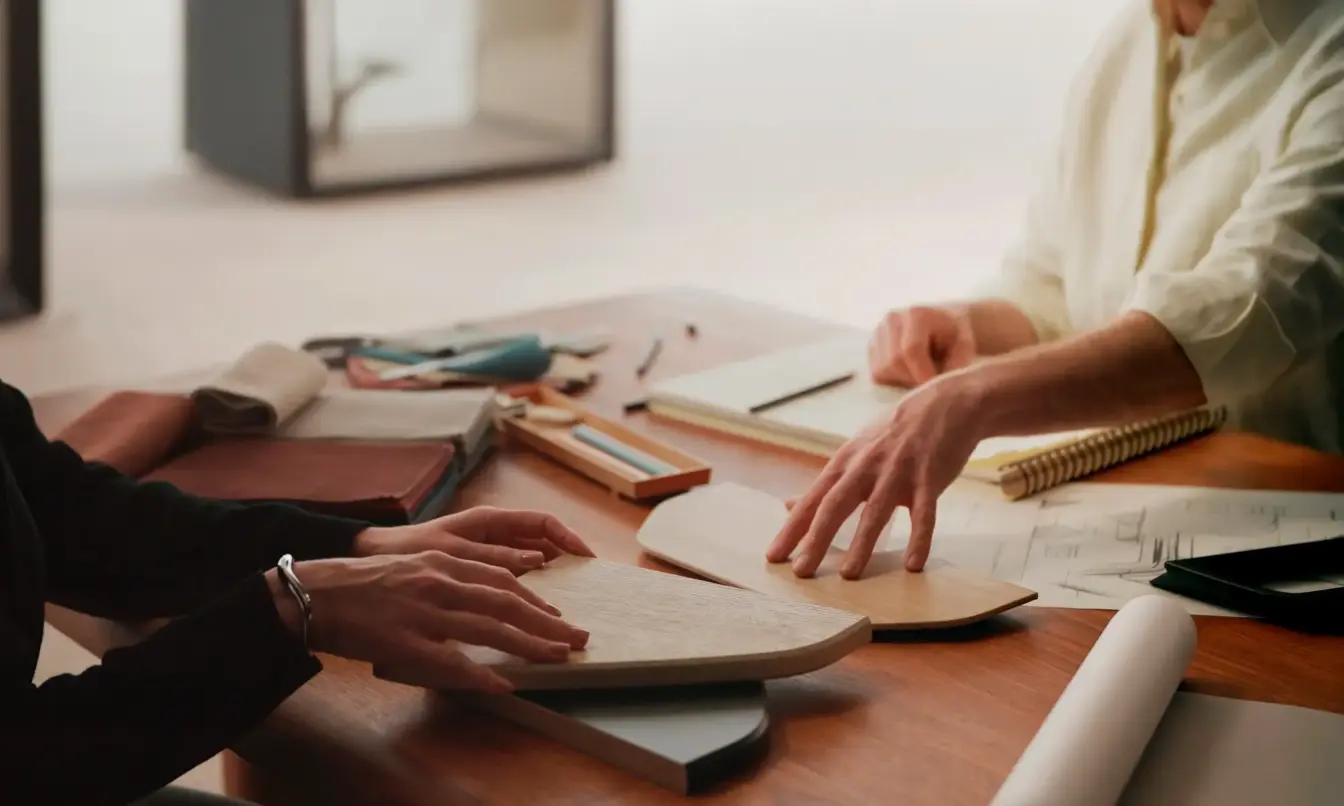 Two people collaborate over documents at a wooden desk with notebooks, rulers, and pens.
