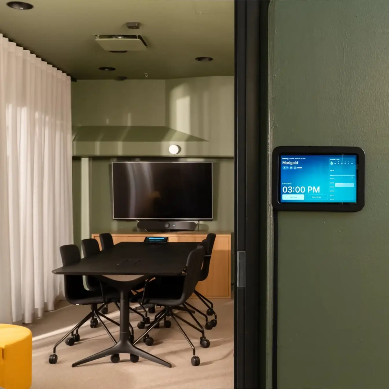 A modern conference room with a dark rectangular table, several black rolling chairs, a wall-mounted screen, and a blue digital schedule display on the green wall.