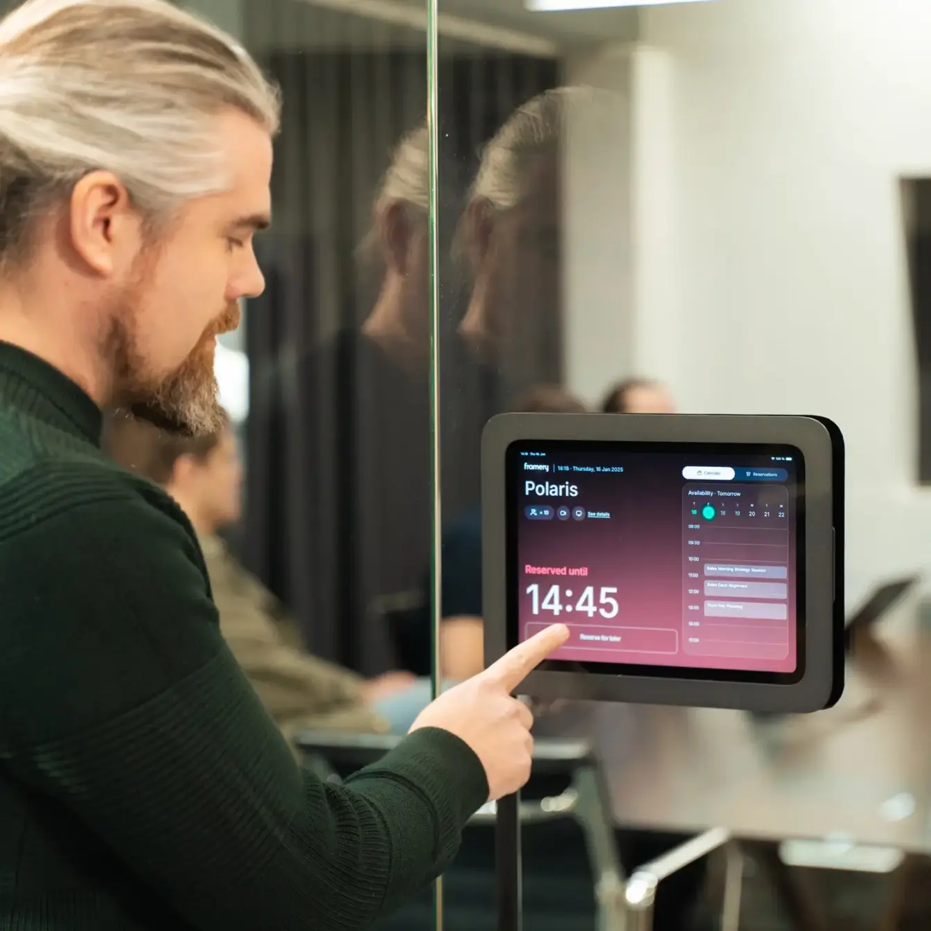 A man with blond hair and a beard in a dark green sweater touches a wall-mounted tablet on a glass wall showing a schedule with the time 14:45.