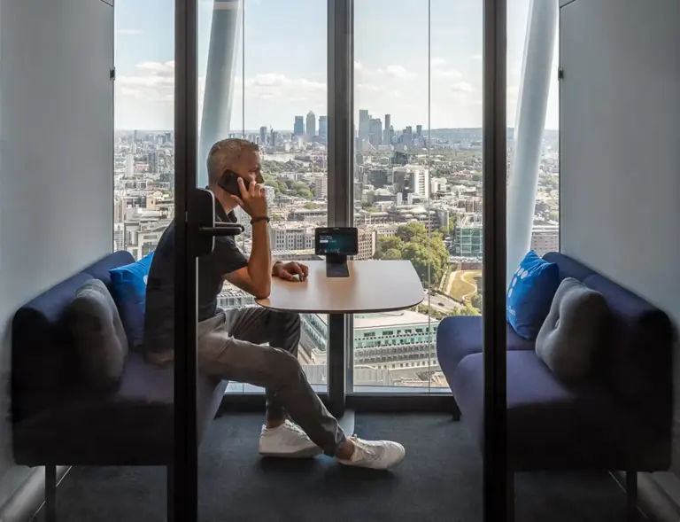 A man sits at a round table in a glass-walled booth with a city skyline view.