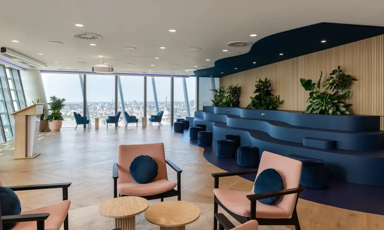 Bright modern lounge with tiered blue seating, pink-cushioned chairs around small round tables, and large windows with plants along the walls.