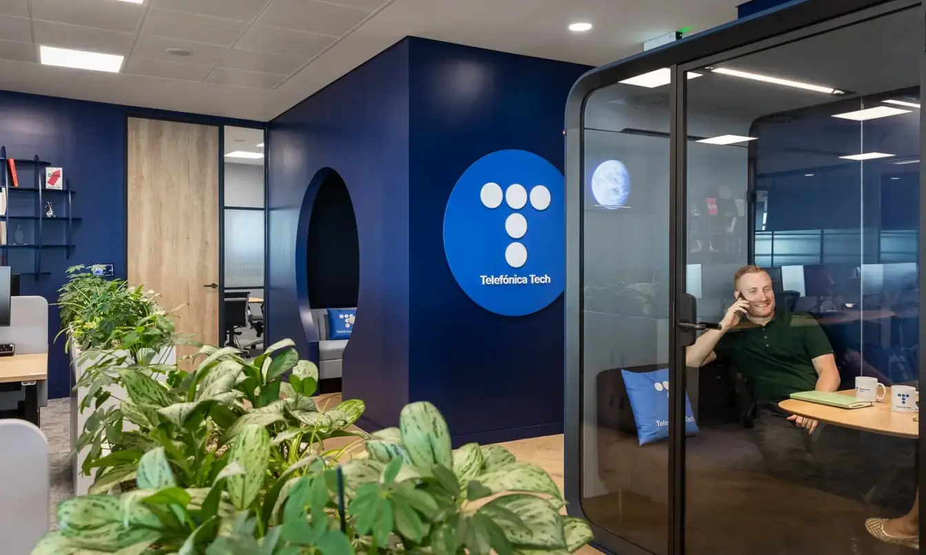 Open-plan office with a blue accent wall and glass-walled meeting room, a person on a call at a table, and green plants in the foreground.