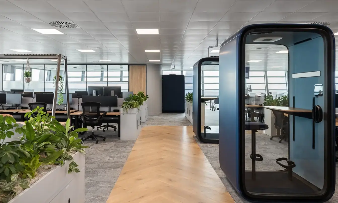 Open-plan office with a long central wooden table, plants along the left, and glass-walled Framery One smart pods on the right.