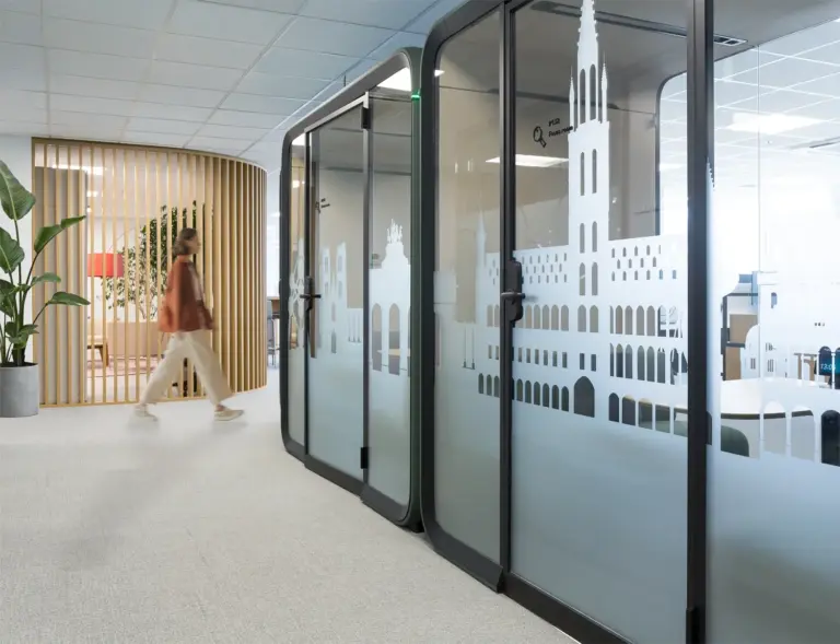 Modern office interior with glass partition walls featuring a white ship motif, a curved wooden wall, and a person walking in the background.