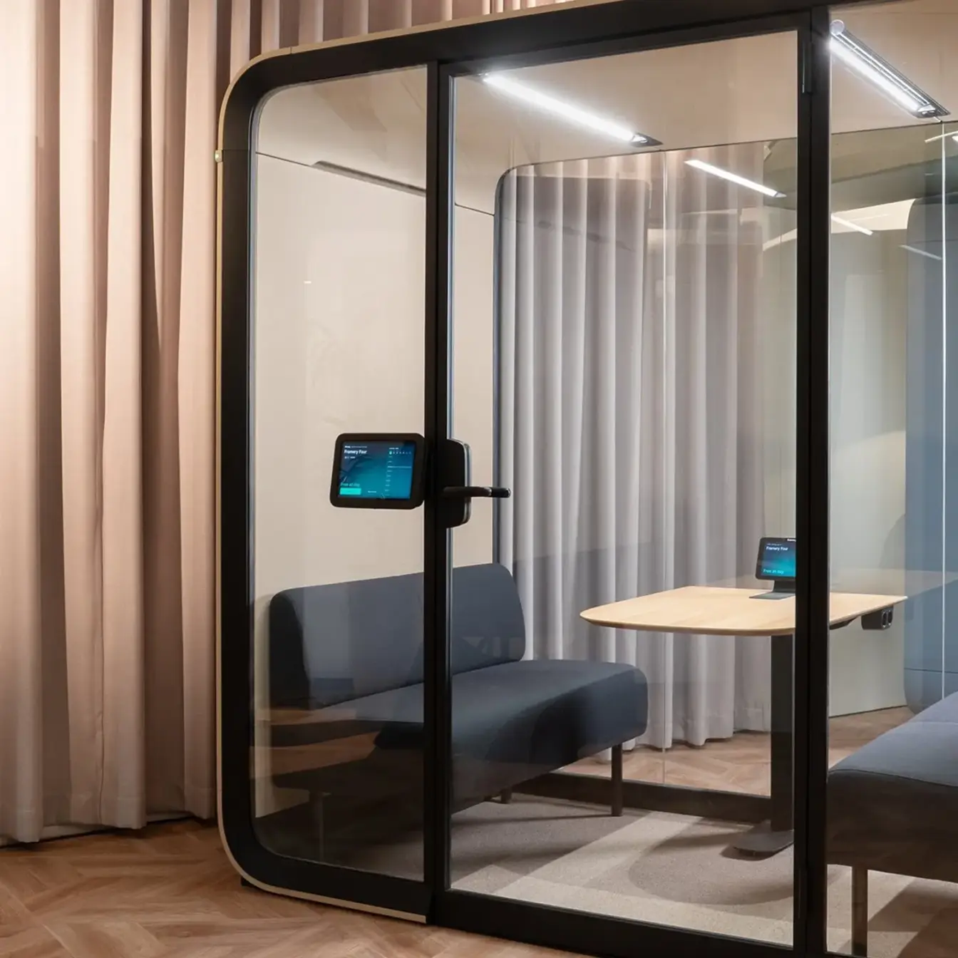 Glass-walled Framery meeting pod with a blue sofa, a small wooden table, and a display.