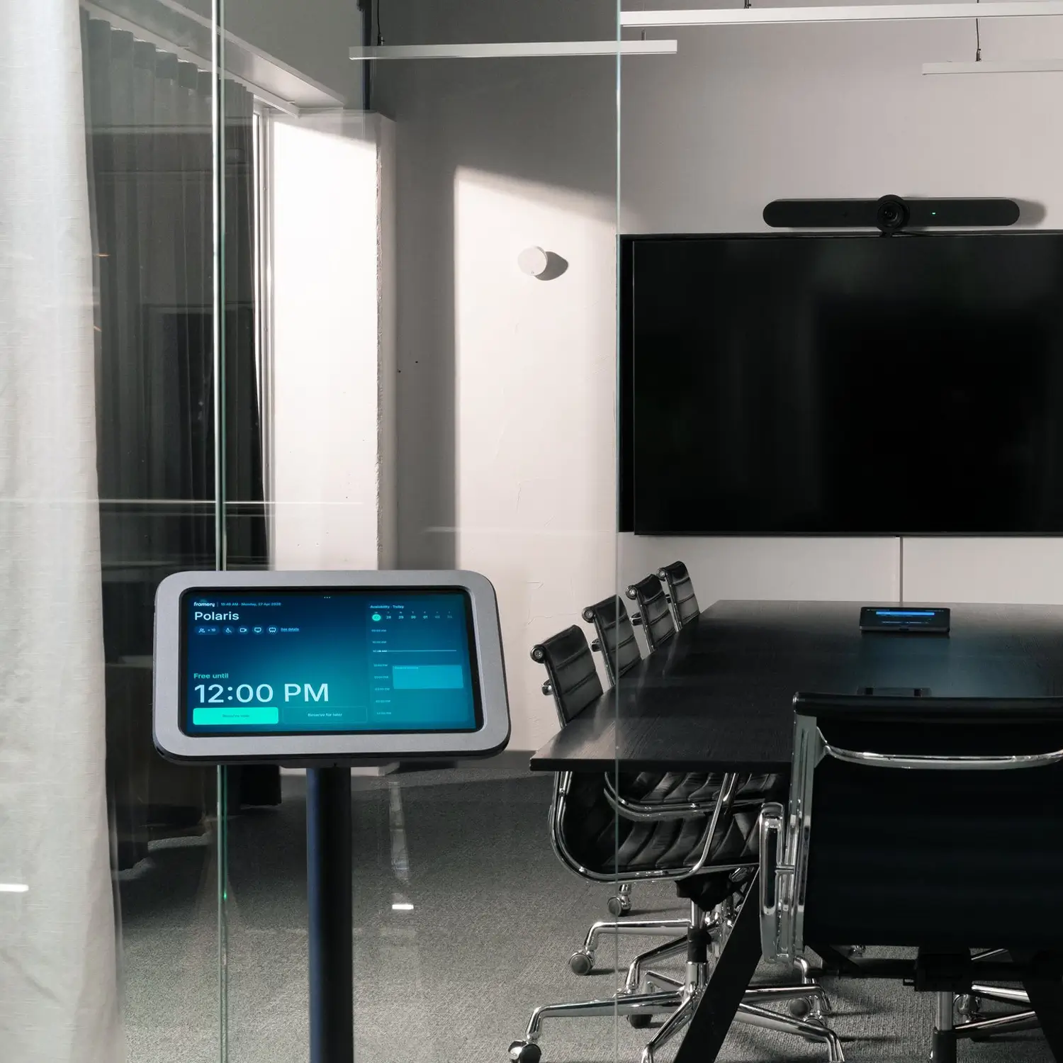 A modern glass-walled conference room with a long table, black chairs, a wall-mounted flat-screen display, and a digital sign in the foreground.