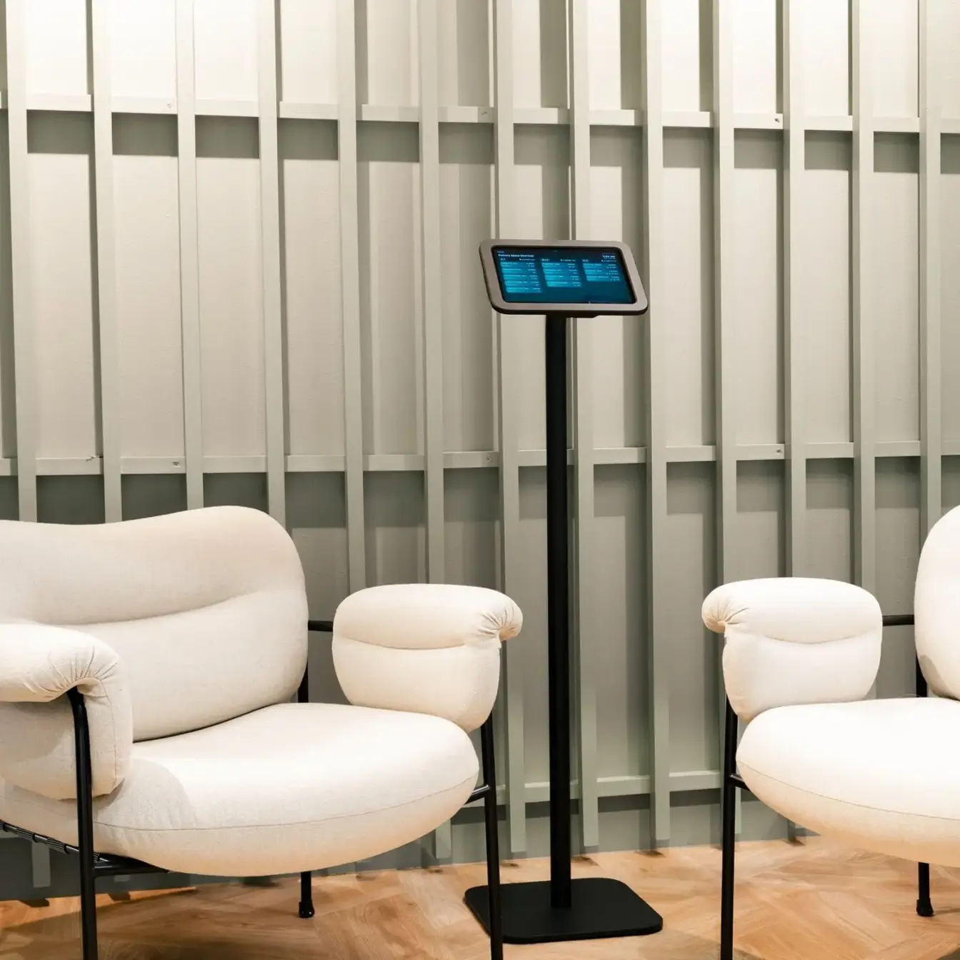 A modern waiting area with white curved chairs, small ottomans, and a tablet on a stand between them.