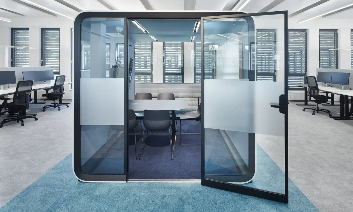Modern open-plan office with a glass-walled Framery Six smart meeting room in the center and desks on both sides.