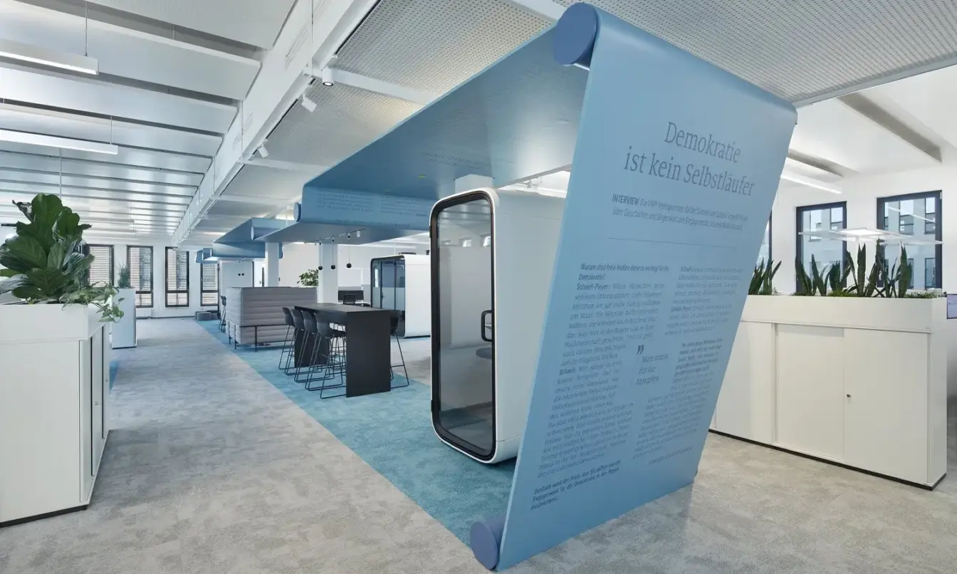 A bright modern open-plan office featuring a blue geometric wall and a Framery One compact pod with a glass door, surrounded by white desks and potted plants.