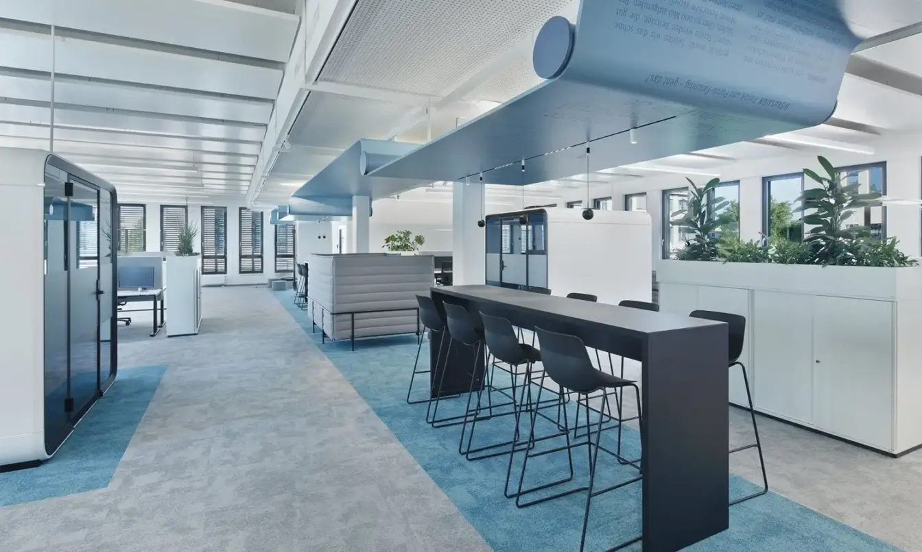 Bright modern open-plan office with white desks, a blue-accent bar-height table, bar stools, white Framery smart office pods and meeting rooms and potted plants.