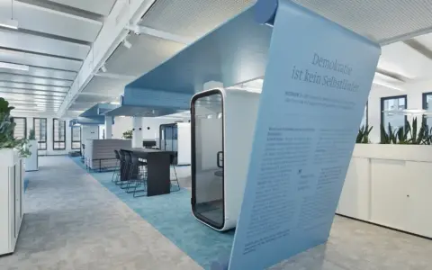 Modern office with a blue curved art installation and Framery office pods, surrounded by desks and plants.