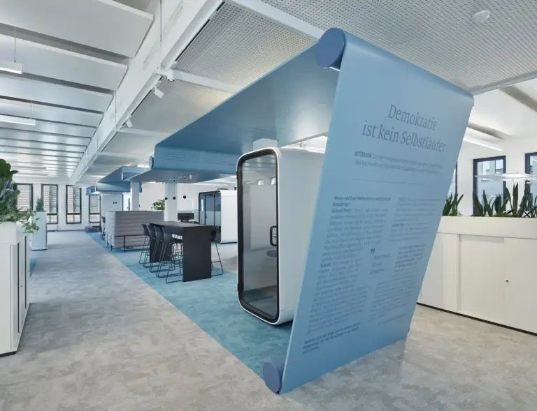 Modern office with a blue curved art installation and Framery office pods, surrounded by desks and plants.