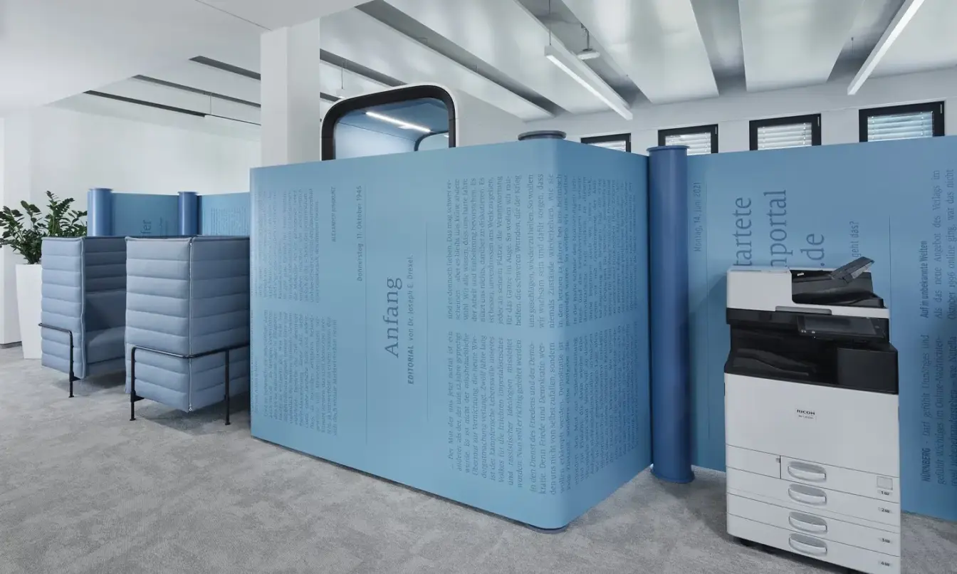 Modern office space with blue modular acoustic partitions, a white four-drawer cabinet with a black printer on top, and plants in the background.