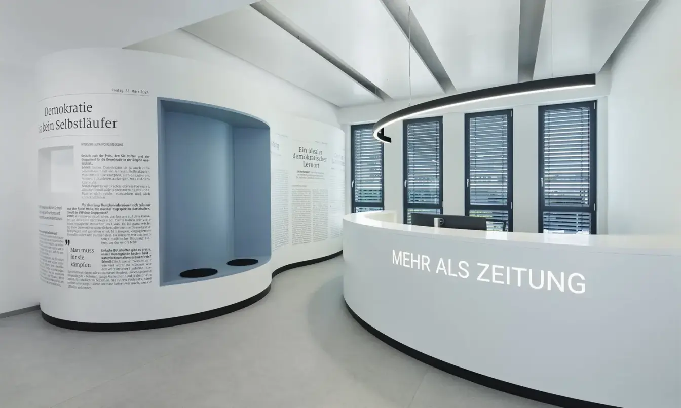 Bright, futuristic office reception featuring a curved white information desk with the text "MEHR ALS ZEITUNG", blue wall niches, and large windows with blinds.