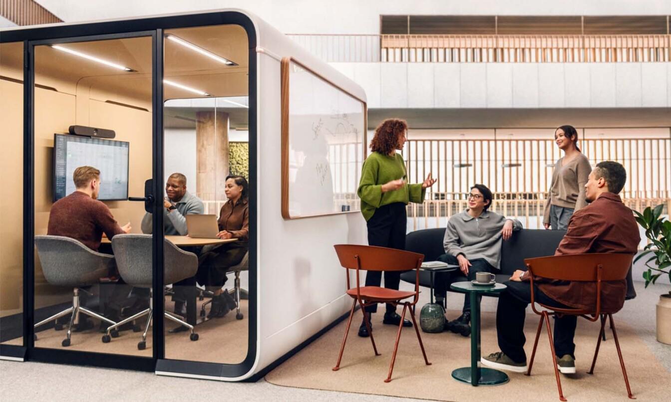 A modern office with a glass-walled meeting room on the left and coworkers chatting around tables on a patio outside.
